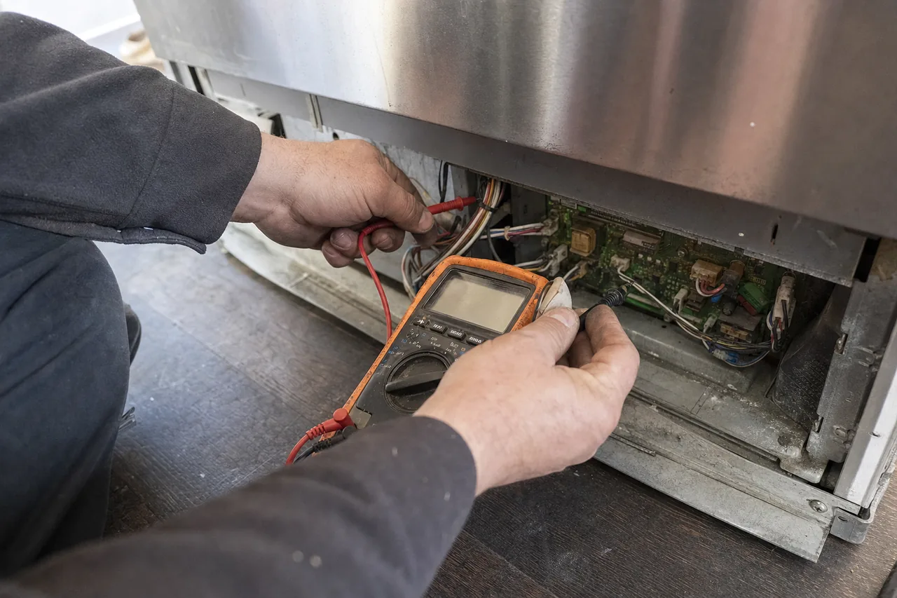 Hands holding an orange-cased multimeter against an exposed circuit board under the base of a refrigerator, ribbon connectors and coloured wiring running away to the right.
