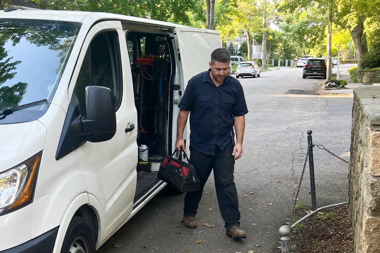 A white service van with its side door slid back on a leafy street, the technician walking up the driveway with his bag, parked cars and mailboxes further along the road.