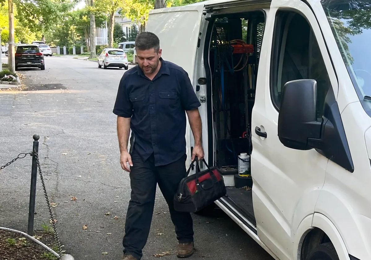 A technician walking up a driveway with a tool bag, the open side of a white van beside him and newer houses visible through the trees ahead.