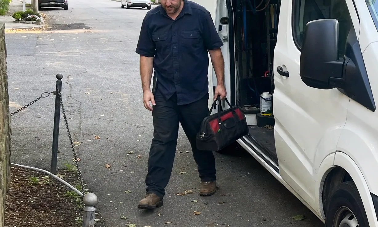 The lower half of a white service van at the kerb with its side door open, the technician’s boots on the asphalt and a leaf-strewn verge alongside.